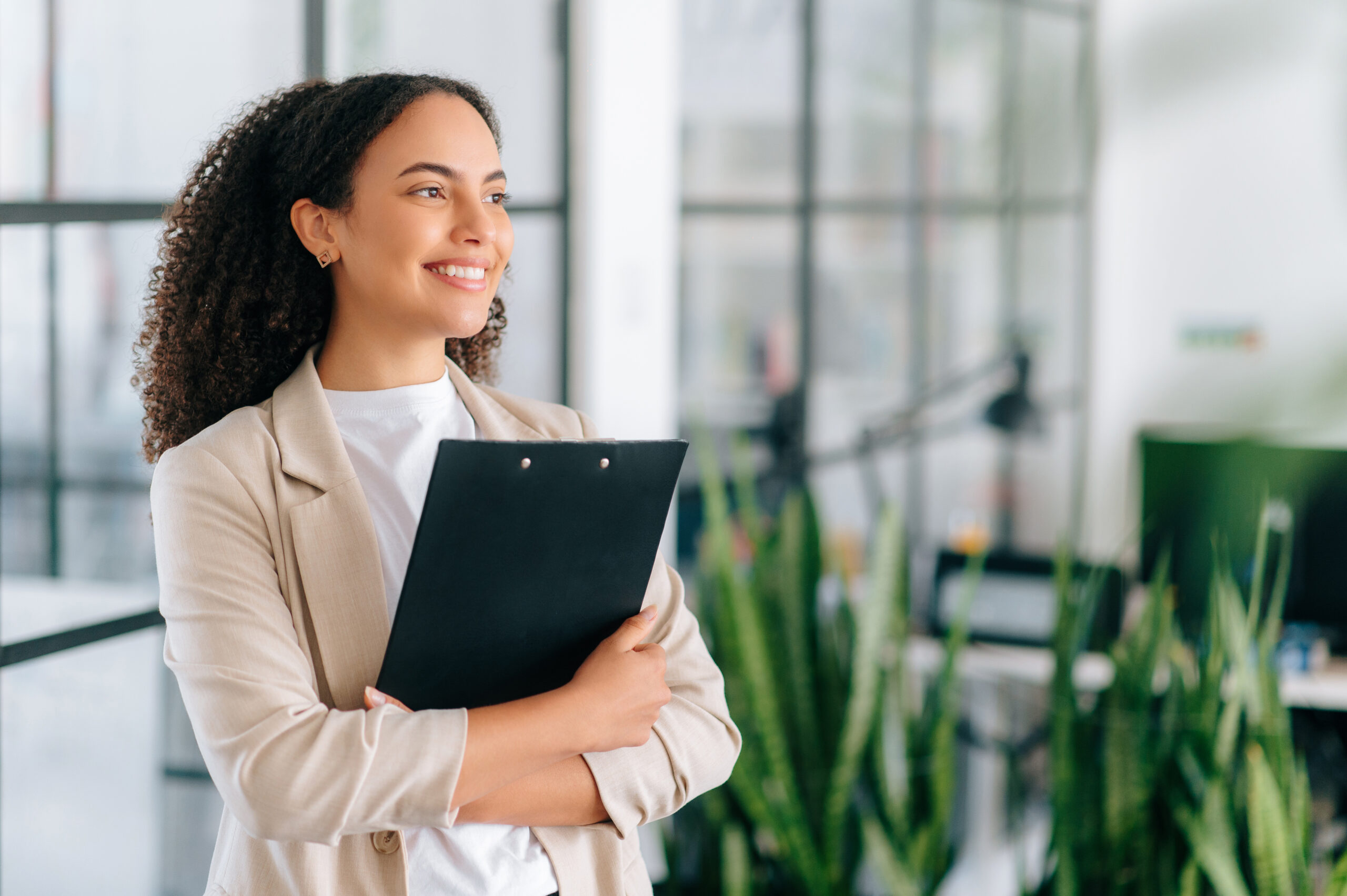 Gorgeous successful Brazilian or Hispanic business woman in elegant clothes, corporate manager, company CEO, secretary, stand in modern office with documents in hands, looks away, smiles